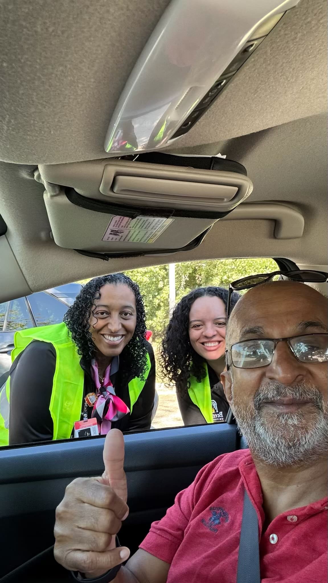A senior driver gives a thumbs up from the driver seat while two volunteers in safety vests smile through the car window.
