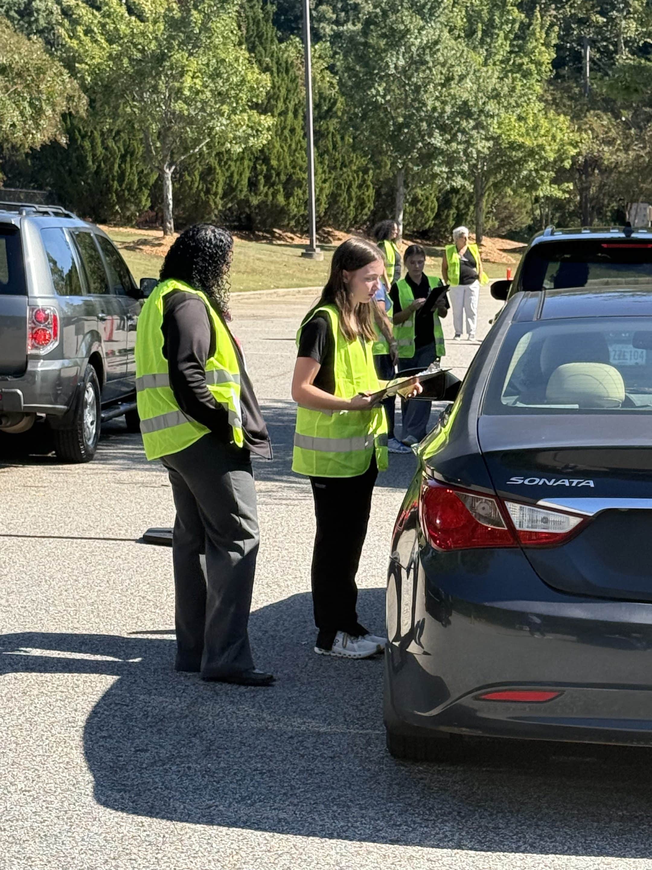Volunteers in safety vests complete a vehicle assessment in the parking lot beside a gray sedan.
